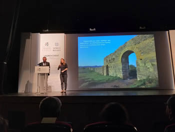 A man and a woman on stage talk while a photograph of ancient walls is projected next to them